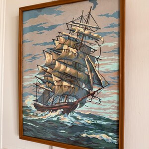 May include: A framed painting of a ship with white sails sailing on a blue ocean with white clouds in the sky.