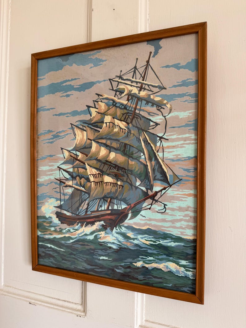 May include: A framed painting of a ship with white sails sailing on a blue ocean with white clouds in the sky.