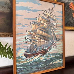 May include: A framed painting of a large sailing ship with white sails against a blue sky and ocean. The ship is in the foreground and the ocean is in the background. The painting is in a wooden frame.