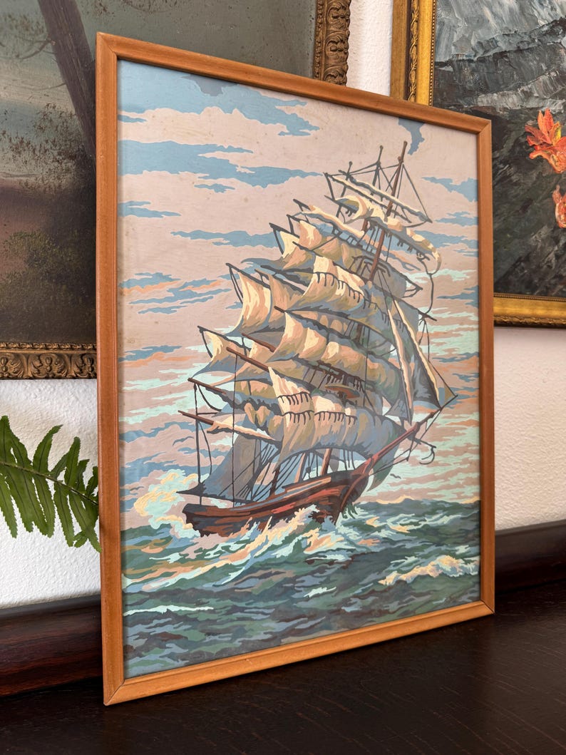 May include: A framed painting of a large sailing ship with white sails against a blue sky and ocean. The ship is in the foreground and the ocean is in the background. The painting is in a wooden frame.
