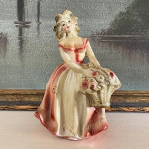 May include: A vintage porcelain figurine of a woman in a long dress, holding a basket of flowers. The figurine is painted in cream, pink, and red hues. The woman's hair is styled in an updo. The figurine is set against a backdrop of a framed painting.