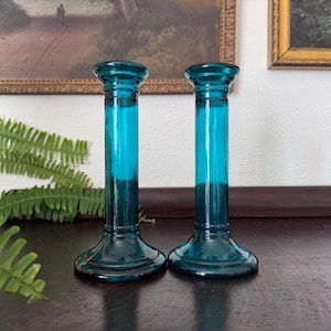 May include: A pair of matching teal blue glass candle holders featuring a cylindrical stem, a flared base, and a circular top rim. The glass is translucent and smooth, with a consistent deep blue-green color throughout the structure of each piece.