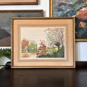 May include: A framed watercolor landscape painting featuring a river scene with a small house and trees in autumn colors. The artwork is in a gold-colored frame and is displayed on a dark wooden surface.
