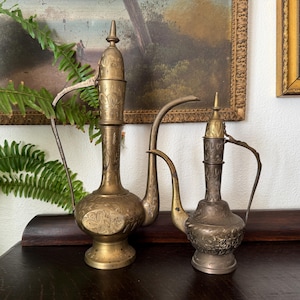 May include: Two antique brass pitchers with ornate floral designs. The taller pitcher has a long spout and a handle, while the smaller one has a shorter spout. Both are on a dark wooden surface, with a painting in the background.