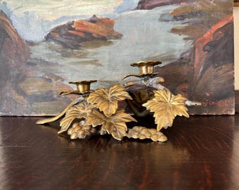May include: A brass double candle holder featuring a detailed botanical design with sculpted grape clusters and large, textured vine leaves. The base supports two circular candle cups, all finished in a warm, metallic gold-toned brass.
