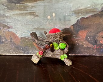 May include: A brown bird figurine perched on a textured tree branch with green leaves and a small red flower. Attached to the branch is a brown nest containing a bright red fabric pincushion, with two silver sewing needles inserted into the cushion.