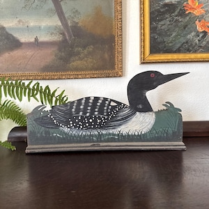 May include: A flat, decorative wooden cutout of a loon bird resting in green grass. The loon features a black head with a red eye, a white neck patch, and a black body with white spotted and striped patterns. It is mounted on a rectangular base.