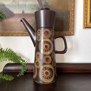 May include: A tall, dark brown ceramic coffee pot featuring a cylindrical body with a tapered top and a matching lid. The exterior is decorated with a repeating pattern of concentric circles in shades of tan and beige, each with a bright orange center.