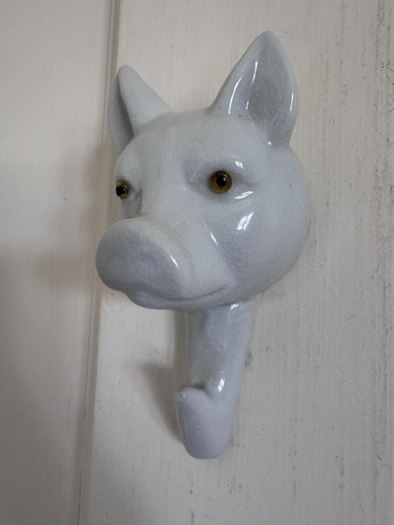 Puede incluir: Un gancho de pared de cer&aacute;mica blanca con forma de cabeza de cerdo. El cerdo presenta un acabado brillante, orejas puntiagudas y dos ojos peque&ntilde;os de color &aacute;mbar oscuro. El hocico sobresale hacia adelante y la parte inferior forma un gancho.