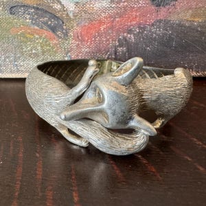 May include: A silver fox bracelet with a detailed design of a fox curled up. The bracelet is made of metal and has a textured finish.