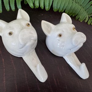 Puede incluir: Un par de ganchos de pared de cer&aacute;mica blanca con forma de cabezas de cerdo. Cada gancho presenta un acabado brillante, orejas puntiagudas, un hocico y peque&ntilde;os ojos oscuros. El gancho se extiende hacia abajo desde la barbilla de cada cerdo.