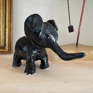May include: A black ceramic elephant figurine with a long trunk. The elephant is standing on a light brown surface.