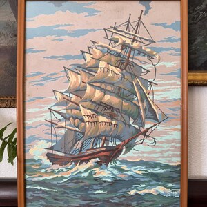 May include: A framed painting of a large sailing ship with white sails, navigating through choppy blue water. The ship is in the foreground, with a cloudy sky in the background.