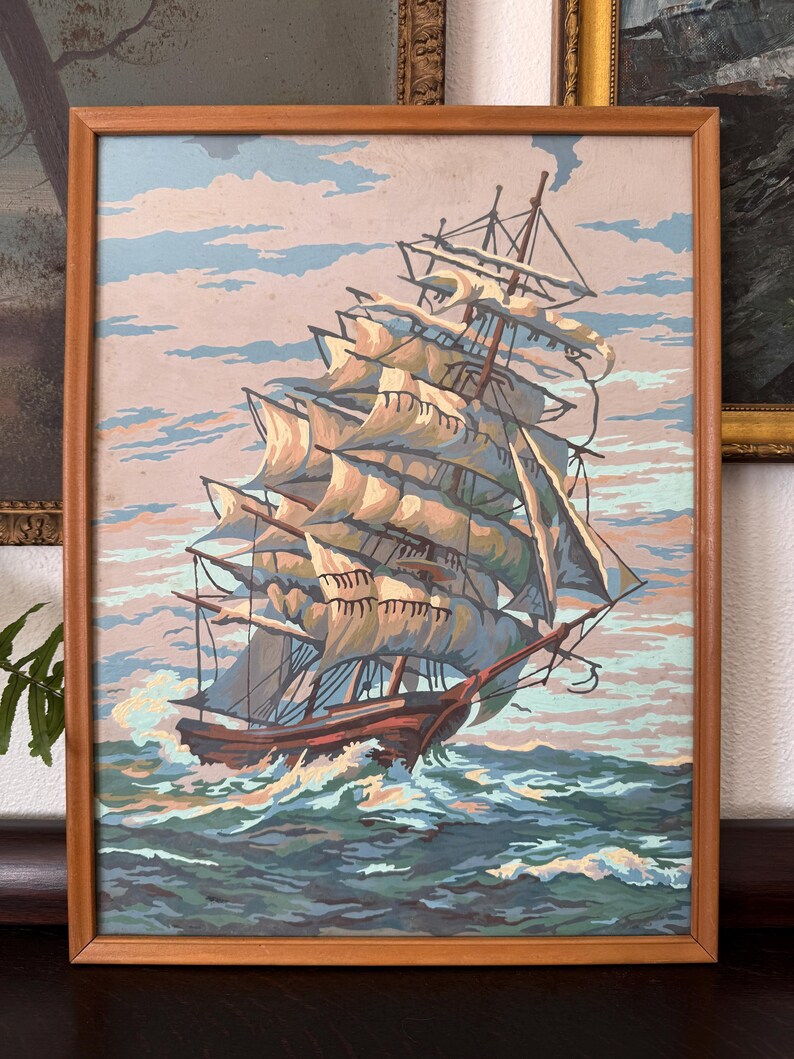 May include: A framed painting of a large sailing ship with white sails, navigating through choppy blue water. The ship is in the foreground, with a cloudy sky in the background.