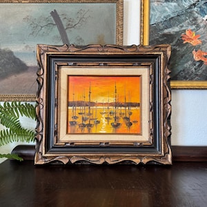 May include: An oil painting of boats in a harbor at sunset, framed in a dark wood frame with ornate carvings. The painting features a warm color palette of orange, yellow, and brown. The painting is displayed on a dark wood surface.