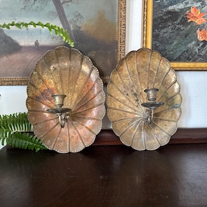 May include: Two antique brass wall sconces. Each sconce features a shell-shaped backplate with a central candle holder. The backplates have a textured, fan-like design and a warm, aged patina. The sconces are mounted on a dark wooden surface.