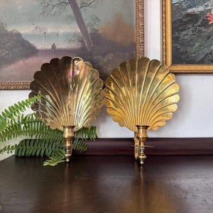 May include: A pair of polished brass wall sconces, each featuring a large, fan-shaped shell backplate with vertical fluted ridges and a single candle holder cup extending from the center base of the shell design for wall mounting.