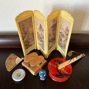 May include: A miniature Japanese room divider screen with a painted landscape scene, a small fan, a pair of wooden sandals, a small box, a doll, and a red and black round box with a painted tree design.