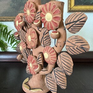 Pu&ograve; includere: Una scultura a candelabro in terracotta a forma di albero, caratterizzata da molteplici fiori piatti e circolari con petali rossi e centri gialli, intervallati da foglie appuntite decorate con strisce nere e piccoli boccioli rotondi su steli.