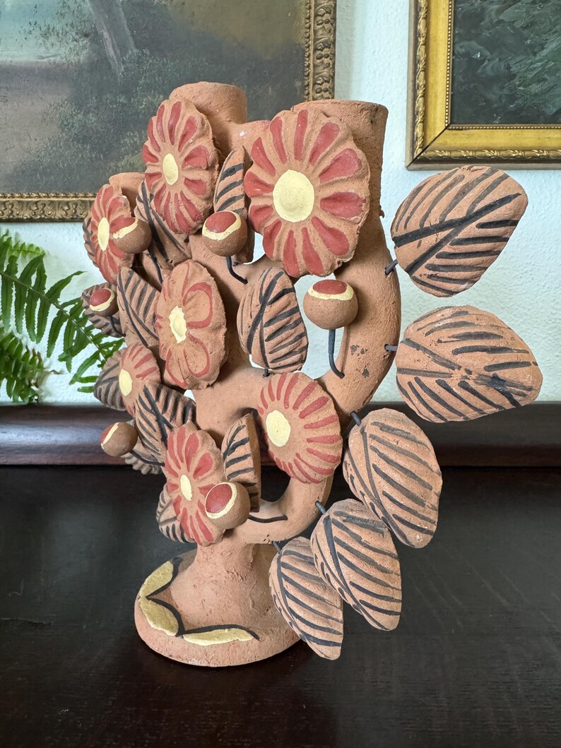 Pu&ograve; includere: Una scultura a candelabro in terracotta a forma di albero, caratterizzata da molteplici fiori piatti e circolari con petali rossi e centri gialli, intervallati da foglie appuntite decorate con strisce nere e piccoli boccioli rotondi su steli.