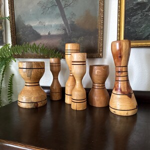 May include: A collection of seven wooden hourglass-shaped decorative objects. Each piece is crafted from light-colored wood with dark bands and varying heights. They are displayed on a dark brown surface, with framed artwork in the background.