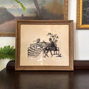 May include: Framed silhouette art depicting a couple seated on a bench, with intricate details. The artwork is in a gold-colored ornate frame. The background is a light beige color. The art is displayed on a dark brown surface.