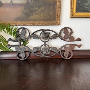 May include: Ornate, dark metal decorative piece featuring a floral design with roses and leaves. The piece is mounted on a dark wooden surface, with a partially rusted appearance on the right side. Decorative art.