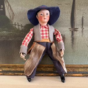 May include: A vintage cowboy doll with a red and white plaid shirt, blue jeans, and a brown fur chaps. The doll is wearing a blue cowboy hat and has a painted face.