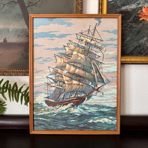 May include: A framed painting of a large, white sailing ship with multiple masts and sails. The ship is sailing on a blue and green ocean with white waves. The painting is in a wooden frame.