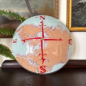 Puede incluir: Una brújula redonda de cristal con un diseño de mapa del mundo. La rosa de los vientos es roja, con los puntos cardinales marcados. El fondo es azul claro con continentes de color beige y líneas rojas.