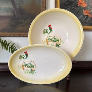 May include: A set of vintage ceramic serving dishes. The set includes a large bowl and an oval platter, both with a cream-colored interior and a yellow rim. Each piece features a rooster and a basket of plants design.
