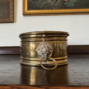 May include: A vintage brass container with a hinged lid. The container features a lion's head with a ring handle on the front. The brass has a warm, aged patina. The container is sitting on a dark wooden surface.