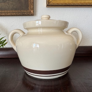 May include: Cream-colored ceramic jar with a lid and two handles. The jar has a dark brown stripe around the base. The lid has a small knob. The jar is sitting on a dark wooden surface.