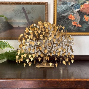 May include: A decorative gold-colored metal tree sculpture with numerous leaves. The tree is displayed on a dark wooden surface, with framed paintings in the background. The sculpture is a unique home decor item.