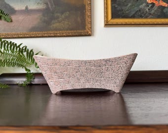May include: A rectangular ceramic bowl with a textured, woven-look exterior pattern in shades of light pink and dark gray. The vessel features a slightly curved top edge and four small, integrated feet at the base, creating a stable, elevated form.