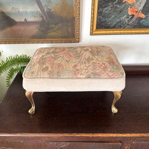 May include: A small, rectangular footstool with a floral patterned top in shades of pink, green, and beige. The sides are a light beige, and the legs are gold-colored, curved, and ornate. The footstool is placed on a dark brown wooden surface.