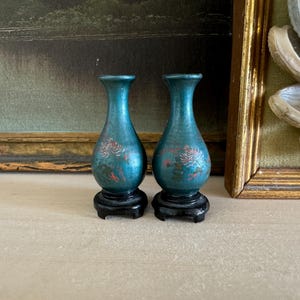 May include: Two small, teal-colored decorative vases with a floral design in red and white. The vases are on black stands. The background includes a framed painting and a framed shell display.