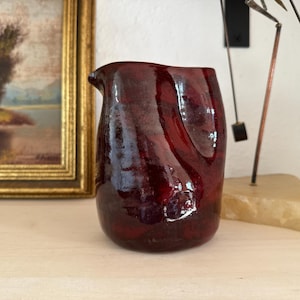 May include: A small, hand-thrown ceramic pitcher with a deep, glossy oxblood red glaze. The vessel features an organic, slightly irregular cylindrical shape with a subtle pouring spout at the rim and a smooth, reflective surface texture throughout.