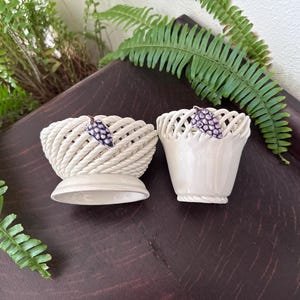 May include: Two cream-colored ceramic bowls featuring a woven lattice design. Each bowl is adorned with a small, purple, sculpted grape cluster accent. The bowls have a smooth, glossy finish and sit on a solid, circular base with a rope-like texture.