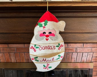 May include: A felt Santa Claus-shaped Christmas decoration with a red hat and white beard. The decoration has two pockets labeled "Santa's Mail" in red script. Green holly accents the design.