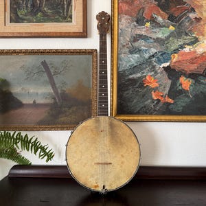 May include: A vintage banjo with a round, aged drumhead and a long, dark wooden neck. The instrument is positioned in front of framed artwork, including landscape paintings. The banjo's design suggests it is an antique musical instrument.