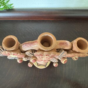 Pu&ograve; includere: Un candelabro in terracotta con tre portacandele cilindrici disposti in fila. La base &egrave; decorata con motivi floreali dipinti a mano nei toni del rosso, giallo e blu, con dettagli a foglia scolpiti che si estendono sui lati.