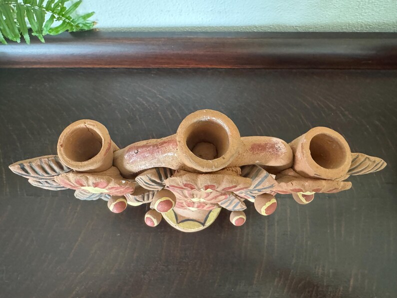 Pu&ograve; includere: Un candelabro in terracotta con tre portacandele cilindrici disposti in fila. La base &egrave; decorata con motivi floreali dipinti a mano nei toni del rosso, giallo e blu, con dettagli a foglia scolpiti che si estendono sui lati.