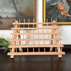 May include: A light-colored wooden thread rack featuring five horizontal tiers supported by two angled side frames. The rack contains numerous evenly spaced vertical pegs designed for organizing spools of thread in a structured display.