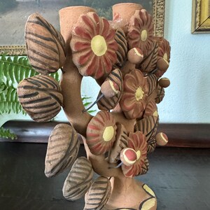 Pu&ograve; includere: Un candelabro in terracotta caratterizzato da una struttura centrale a rami decorata con fiori e foglie scolpiti. I fiori presentano petali rossi e centri gialli, mentre le foglie sono caratterizzate da una texture a strisce marrone scuro.