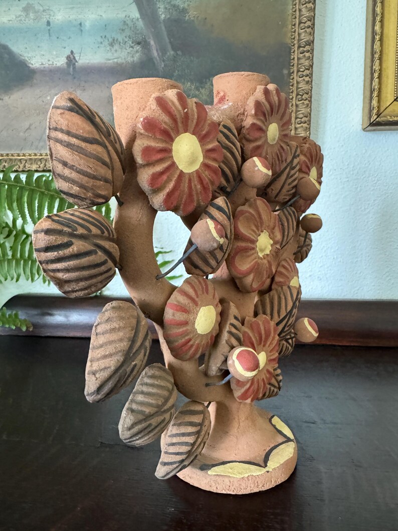 Pu&ograve; includere: Un candelabro in terracotta caratterizzato da una struttura centrale a rami decorata con fiori e foglie scolpiti. I fiori presentano petali rossi e centri gialli, mentre le foglie sono caratterizzate da una texture a strisce marrone scuro.