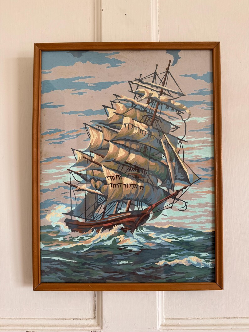 May include: A framed painting of a large sailing ship with white sails against a blue and gray sky. The ship is sailing on a blue and green ocean with white waves.