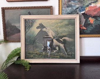 May include: A framed print featuring a black horse and a white horse running through a grassy field under a stormy sky with lightning. The artwork is enclosed in a light-colored wooden frame with visible signs of wear and aging.