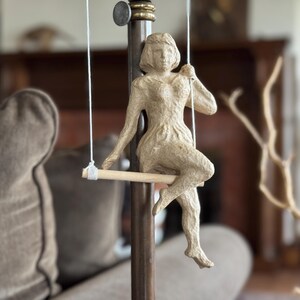 May include: A beige paper sculpture of a woman seated on a swing. The figure is suspended by white strings from a wooden bar. The sculpture is attached to a dark brown pole, with a blurred background of a living room.