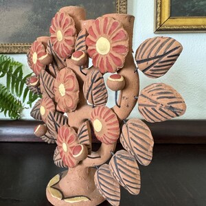Pu&ograve; includere: Un candelabro in terracotta a forma di albero, caratterizzato da fiori dipinti a mano in rosso e giallo, foglie a righe marrone scuro e piccoli boccioli rotondi. La scultura ha una base circolare e due portacandele in cima, finitura opaca.